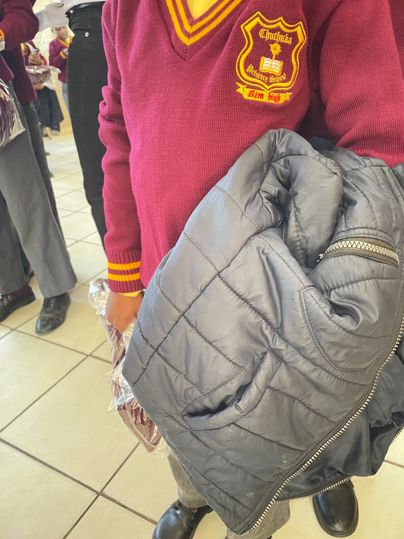 Close-up of the Thuthuka Primary School crest and its Aim High motto on a maroon school jersey, 30 July 2026